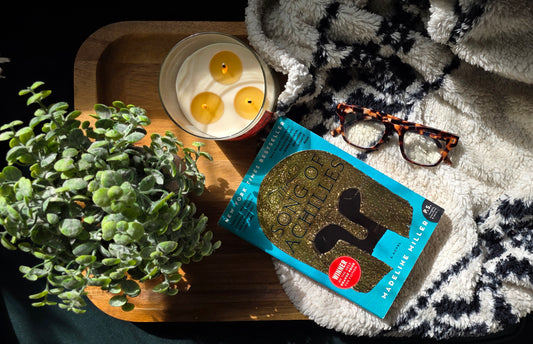 Book titled 'The Song of Achilles' by Madeline Miller on a blanket with a candle and plant in the background.