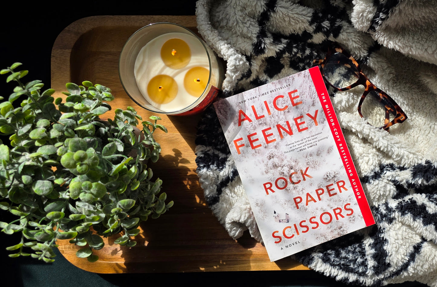 Book titled 'Rock Paper Scissors' by Alice Feeney on a wooden tray with a candle and plant.